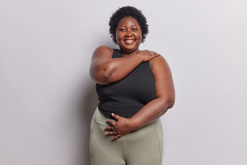 Wall Mural - Photo of dark skinned overweight woman embraces herself has fat body dressed in black t shirt and leggings smiles broadly isolated over white background. Plump African female wants to loose weight