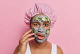 Fototapeta  - Photo of young man examines his skin applies beauty clay mask with cucumber slices on face for rejuvenation and skin regeneration wears bath hat and white t shirt isolated over pink studio wall