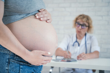 Wall Mural - Doctor obstetrician gynecologist at his desk in the background. Close-up of a pregnant woman's belly.