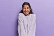 © WHstudio Leushin N - Waist up shot of positive Iranian woman with dark hair smiles happily keeps eyes closed recalls something funny dressed casually isolated over purple background. Positive human emotions concept
