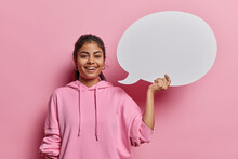 Positive Lovely Iranian Woman With Dark Hair Smiles Gladfully Holds Blank Speech Bubble Gives Promotional Opportunity Smiles Gladfully Wears Casual Sweatshirt Poses Against Pink Studio Background.