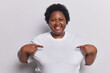 Clothing template concept. Positive overweight African woman with curly hair points at copy space of t shirt suggests to write you logo here isolated over white background. Place you test here