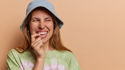 Wall Mural - Horizontal shot of positive European woman bites finger and looks aside smiles gladfully has curious expression notices something funny wears panama and t shirt isolated over brown background