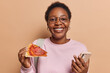 © WHstudio Leushin N - Good looking dark skinned African woman eats delicious pizza enjoys eating fast food using mobile phone leaves positive feedback to delivery service smiles gladfully poses with rucksack indoors
