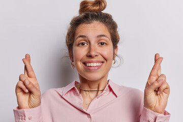 Wall Mural - Superstitious European woman with hair gathered in bun crossing fingers praying for miracle hoping for good luck wears pink shirt smiles toothily isolated white background. Make wish come true