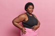 Sideways shot of curly haired African woman keeps hands on stomach and hip has serious expression concentrated at camera isolated over pink background. Overweight dark skinned female in sportswear