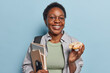 Smiling dark skinned teenage girl with short curly hair eats glazed doughnut holds notepads with smartphone has delicious snack wears spectacles and casual clothing isolated over blue background.