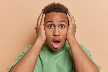 Wall Mural - People emotions concept. Indoor photo of young confused African american guy holding head with hands looking straight at camera standing in centre isolated on beige background not knowing what to do