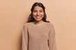 Portrait of pleasant looking Iranian girl with toothy smile has good mood dressed in casual knitted jumper looks directly at camera isolated over brown background. People and positive emotions concept
