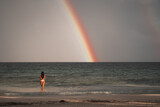Fototapeta Tęcza - A girl in a bathing suit stands on the beach in front of a rainbow.