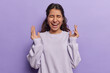 Waist up shot of overjoyed Iranian woman with dark hair exclaims loudly keeps hands raised and laughs at something dressed in casual sweatshirt isolated on purple background. People happiness concept
