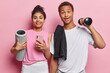 Sporty concept. Indoor shot of young glad happy smiling European female and African american male holding massage roller and dumbbell ready for training to be healthy and keep fit on pink background