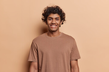 portrait of happy hindu man with curly hair smile toothily keeps arms down dressed in casual t shirt