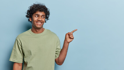 Wall Mural - People positive emotions concept. Studio waist up of young happy smiling Hindu male standing on left isolated on blue background pointing at blank space for your promotion wearing olive t shirt