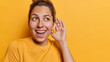 Horizontal shot of healthy young cheerful woman keeps hand near ear tries to overhear rumors listens private information with interest has curious expression isolated over yellow background.