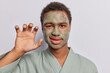 Horizontal shot of African man with curly hair makes paw with hand smirks face applies natural clay mask for glowing skin wears domestic robe isolated over white background. Beauty procedures at home