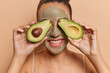 Horizontal shot of cheerful unrecognizable smiling woman covers eyes with two avocado pieces takes care of facial skin uses natural organic cosmetics isolated over brown background. Beauty concept