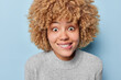 Portrait of surprised curly haired woman bites lips and stares with widely opened eye has curious expression wears casual grey jumper isolated over blue background. Human facial expressions concept