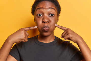 People emotions concept. Indoor close up of young African american model with rounded lips wearing black t shirt standing isolated on yellow background keeping hands raised pointing at cheeks