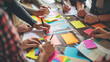 © Studio Nova - ?lose-up view of several hands collaboratively working on a project with colorful sticky notes, papers, and digital devices on a wooden table.