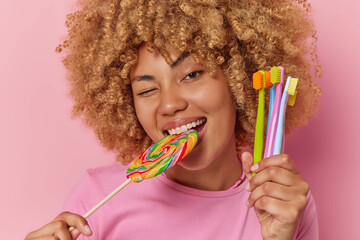 Wall Mural - Dental care concept. Studio close up of young pretty glad smiling African american female eating delicious lollipop holding toothbrushes standing in centre isolated on pink background wearing t shirt