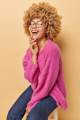 Wall Mural - Vertical shot of overjoyed curly haired woman laughs joyfully as hears something funny poses on chair being in good mood wears spectacles pink jumper and jeans isolated over brown background
