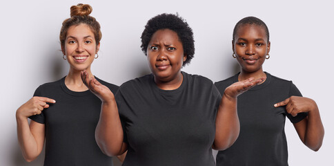 Wall Mural - Photo of mixed race women show mock up space on t shirt smile gladfully advertise something. Dark skinned hesitant overweight African woman spreads palms cannot decide feels hesitant. Human perception