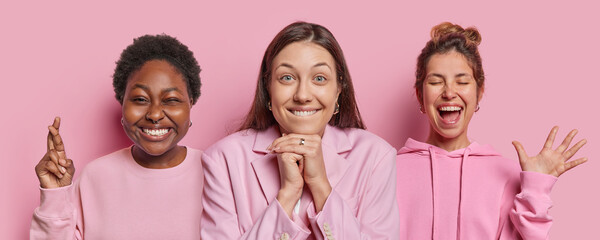 Wall Mural - Photo of three emotional women dressed in casual clothes smile gladfully keep fingers crossed palms raised up smile positively make wish exclaim loudly isolated over pink background. Emotions concept