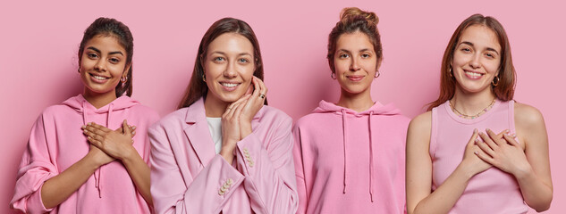 Wall Mural - Horizontal shot of four mixed race young women smile gladfully press hands to heart make gratitude gesture have happy expressions express positive emotions stand in row against pink background