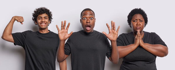 Wall Mural - Photo of shocked scared dark skinned man stares amazed keeps palms raised up cheerful Hindu man shows biceps displeased overweight African woman begs for something wear black t shirts stand indoor