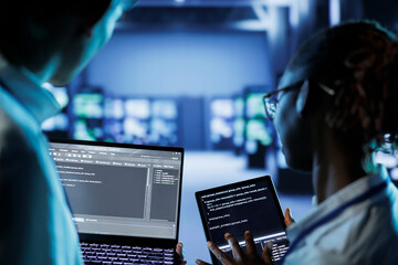 IT developers building firewall systems in server room housing advanced infrastructure. Licensed specialists running code on tablet and laptop, to upgrade data center rigs handling demanding workloads
