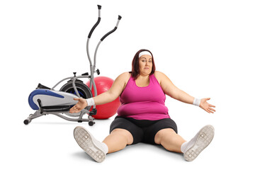 Wall Mural - Tired plus size woman in sportswear sitting on the ground in front of a ball and stationary bike