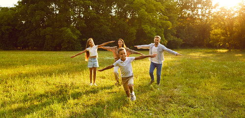 Wall Mural - Happy family running and 'playing plane having fun in summer park enjoying sunny day in nature. Mother, father and their children boy and girl walking on green grass outdoors at sunset together.