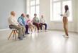 © Studio Romantic - Young woman speaking on meeting in nursing home in front of a small group of senior people about dementia prevention. Elderly men and women sitting on chairs in row and listening speaker on seminar.
