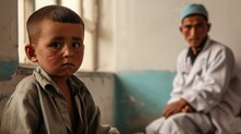 A Little Boy Sits With Sad Eyes In A Hospital Bed Against The Background Of An Old Peeling Wall, In The Background Is A Blurred Background Of A Doctor In A White Coat. Health And Help Concept