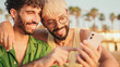© Andrii Nekrasov - Close-up of young gay couple sitting embracing, smiling and laughing have fun with mobile phone while sitting on the beach