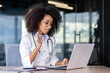 © Liubomir - African american young female doctor sitting in clinic office at table and talking online with patient, consulting and greeting with hand to camera