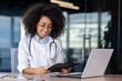 Smiling young African American female doctor working in clinic office with laptop and documents, sitting at table, holding folder, reading, studying, analyzing