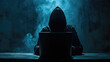 Silhouette of a person in a hoodie working on a laptop in a dark room, with a blue light emanating from the screen.