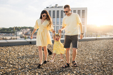 Lovely man and woman holding hands of little beautiful girl and lifting her into air outdoors. Smiling family wearing matching yellow outfits creating happy childhood to cute daughter on rooftop.