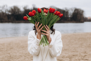 Wall Mural - Attractive brunette woman walking on the beach shore in moody cloudy windy weather with bouquet of red tulips flowers, dressed in white suit jacket. International Women`s Day 8th March concept