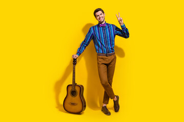 Sticker - Full size photo of positive guy dressed striped shirt stand with acoustic guitar showing v-sign isolated on yellow color background