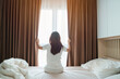 © Jo Panuwat D - happy woman stretching on bed after wake up, young adult female rising arms and looking to window in the early morning. fresh relax and have a nice day concepts
