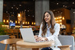 © Graphicroyalty - Young woman, digital nomad working remotely from a cafe, drinking coffee and using laptop, smiling.