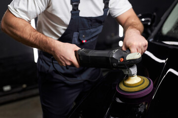 Wall Mural - Cropped professional car service male worker with orbital polisher, polishing black luxury car hood in auto repair shop. Close-up hands