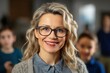 © Vilaysack - A cheerful senior female teacher with glasses standing in a classroom, with young students visible in the background.