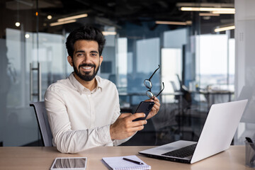 Wall Mural - Portrait of a smiling young Indian man sitting in the office at the desk, holding glasses and a mobile phone, looking confidently at the camera