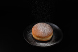 Fototapeta  - Single fresh baked donut berliner sprinkled with falling sugar powder on retro dark plate on black background. Sweet doughnut closeup.