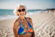 Happy old senior woman in bikini with wrinkled face, flabby tanning skin and gray hair enjoys retirement on sea beach, mature granny in sunglasses and swimwear, elderly model