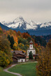 Bavarian Alps with church of Wamberg in Garmisch-Partenkirchen during autumn, snow-covered mountains in the background, dramatic cloudy sky, colored leaves and trees and road in foreground